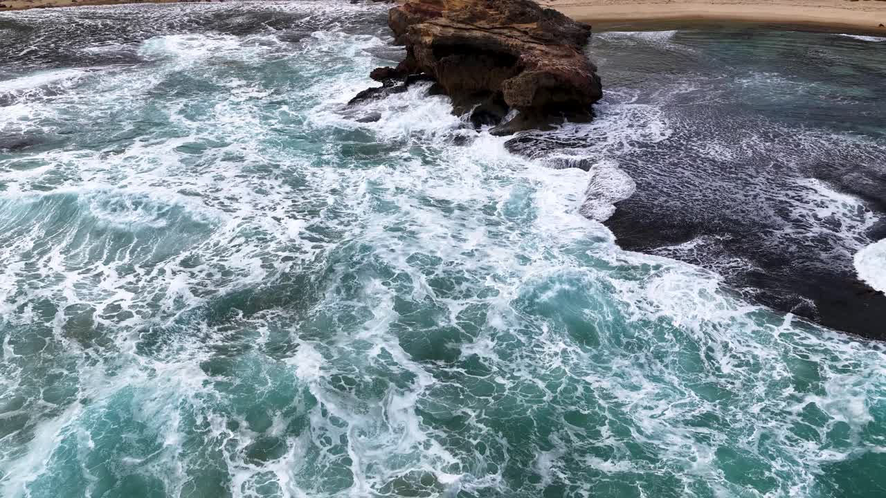 Drone captures turquoise waves breaking against rugged coastal rock formation under natural daylight at Portsea