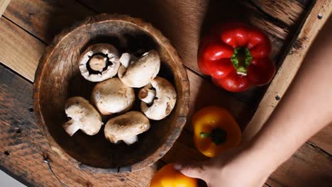 hand putting two yellow pepper next to a red pepper and wooden bowl with white mushrooms inside on rustic wooden texture as background-flat lay, steady cam