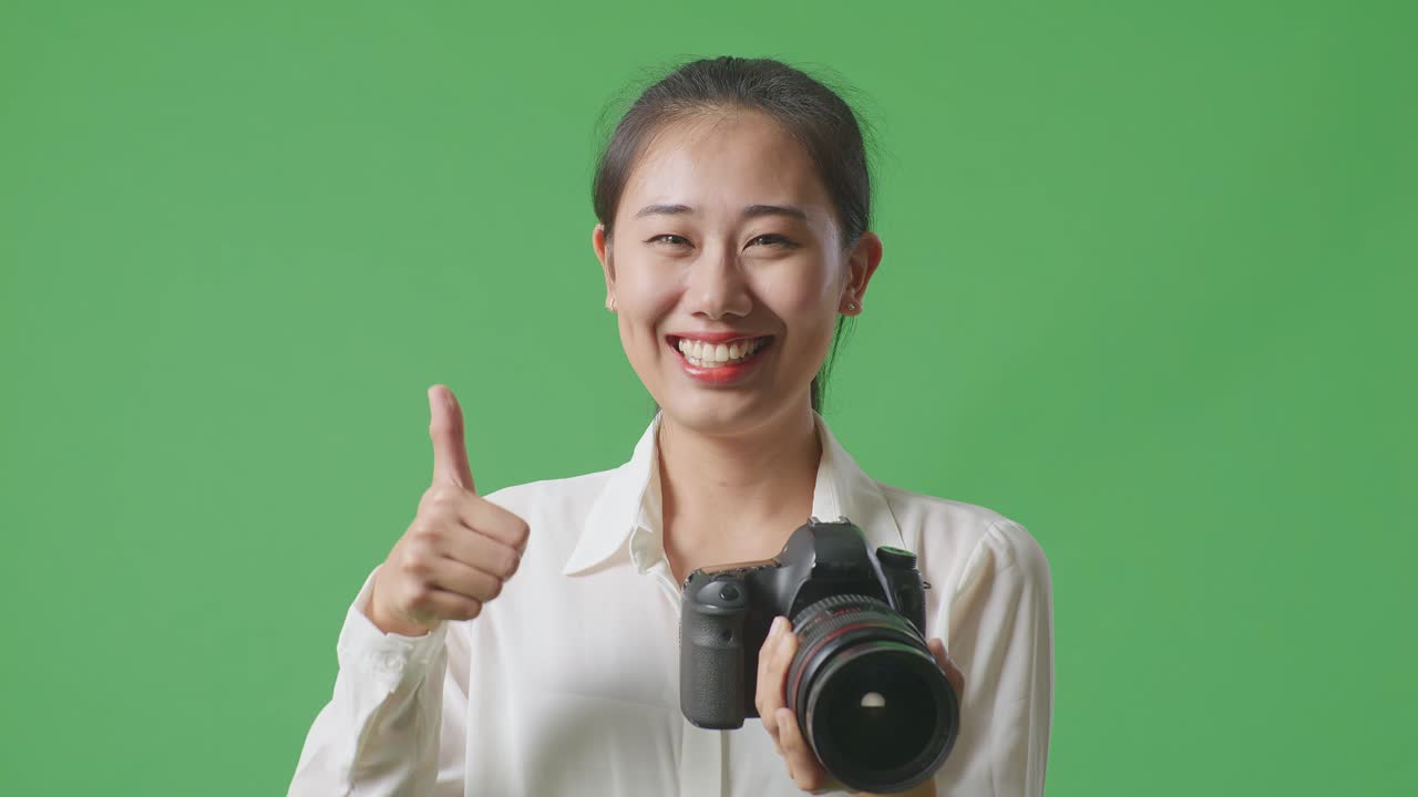 스튜디오에서 녹색 스크린 배경에 서 있는 동안 엄지손가락을 올려놓고 미소 짓는 카메라를 가진 아시아 사진작가의 클로즈업
