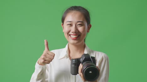 close up of asian photographer with a camera showing thumbs up gesture and smiling while standing on green screen background in the studio