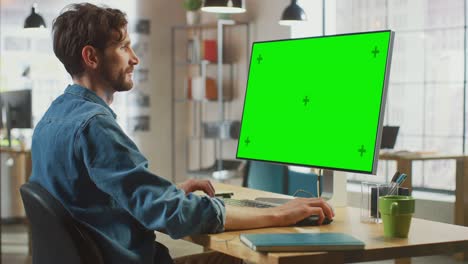 el diseñador creativo masculino con barba y camisa de vaqueros trabaja en su computadora personal con una gran pantalla verde. trabaja en un cool loft de oficina. otra compañera creativa camina en el fondo.