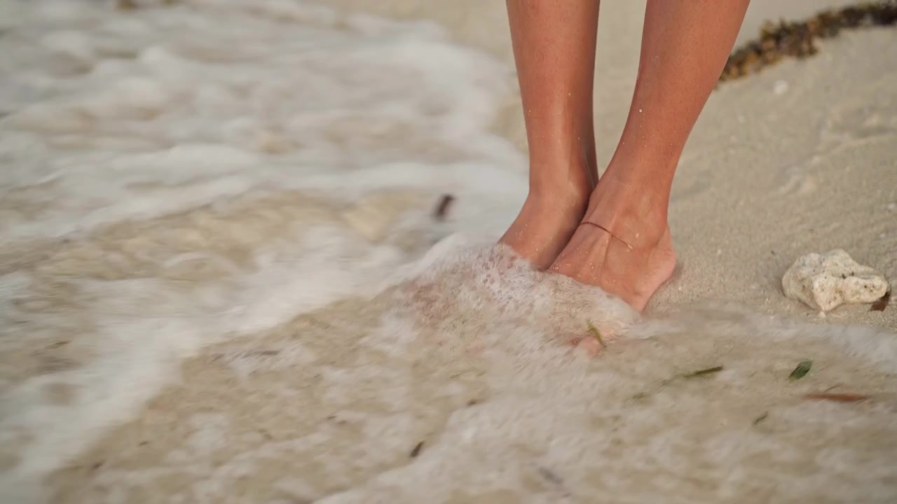 Piedi su una calda spiaggia sabbiosa, onde delicate che accarezzano le dita dei piedi, orme sulla sabbia di un viaggiatore rilassato che esplora la riva con conchiglie sparse, ciottoli e legni alla deriva, luce soffusa del tramonto e schiuma di marea calma