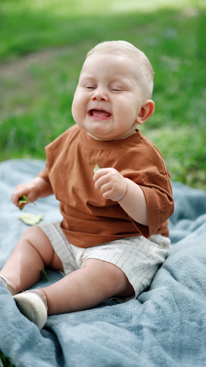 Adorable chubby Babyboy with blond hair sits on the grass outdoors. Blue-eyed kid sneezes funnily. Vertical video.