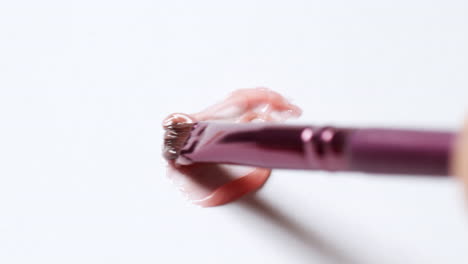 primer plano de cepillado de un poco de bálsamo labial rosa melocotón, usando un aplicador de cepillo labial en una tabla blanca y limpia