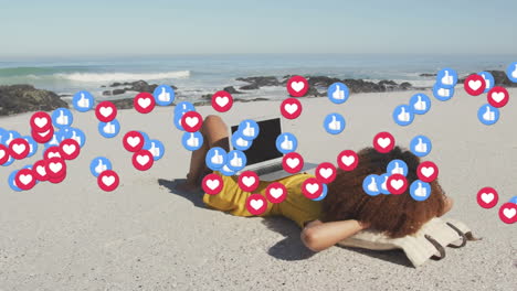 animation of red heart love and like digital icons over woman using laptop on beach