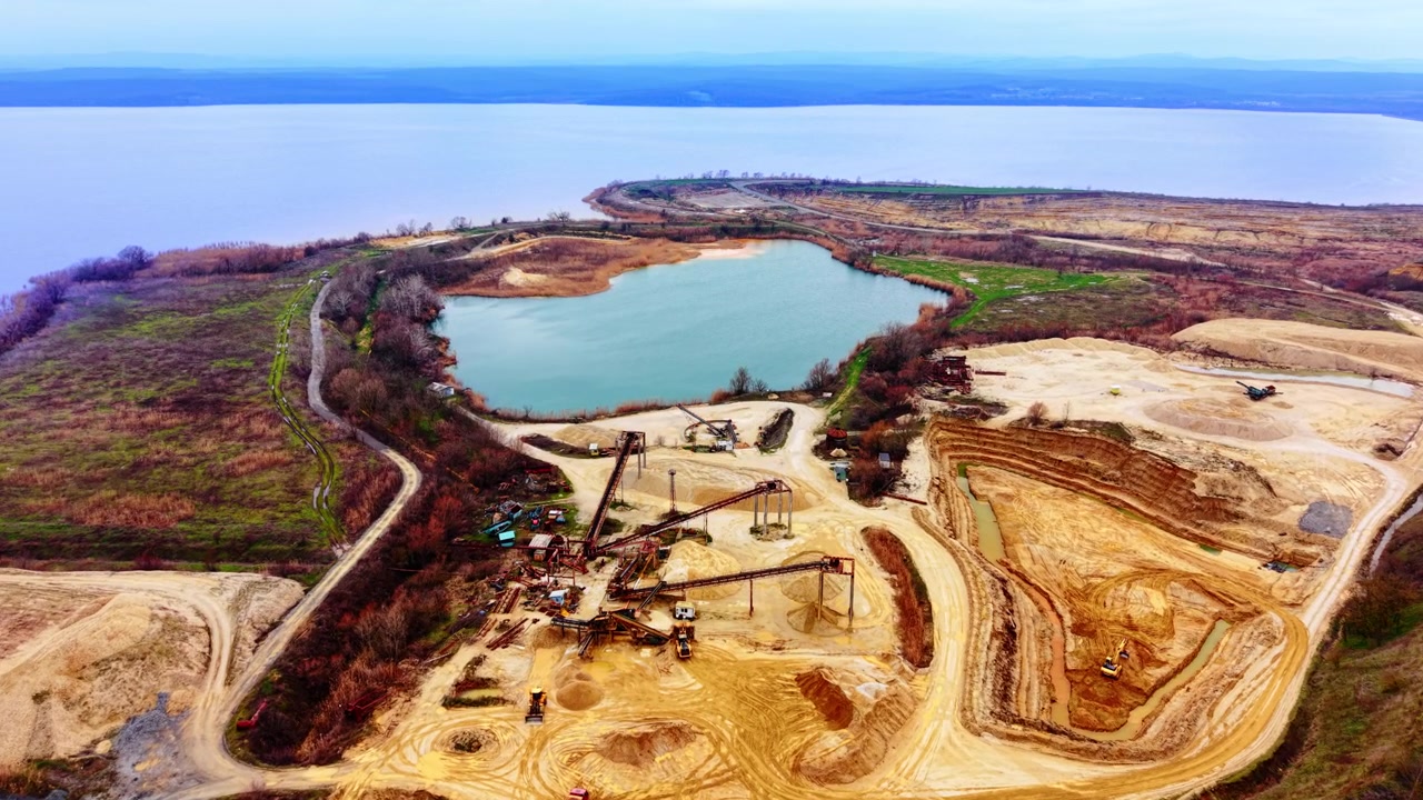 Aerial view of sand quarry with conveyor systems near a lake, Bulgaria. Drone perspective showing extraction plant, sandy terrain, teal pond and distant river under hazy sky.