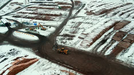 Aerial view of wheel loader with grapple bucket driving through large piles of logs covered in snow at Canadian sawmill