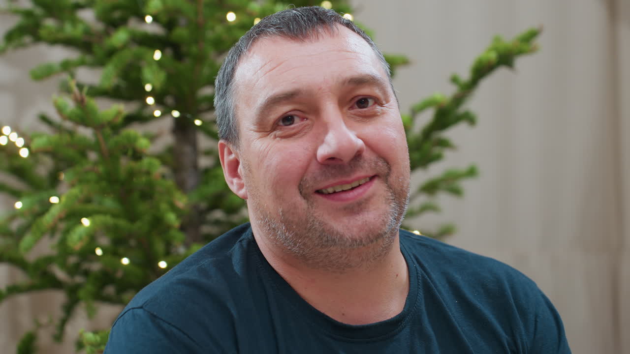Portrait view of cheerful middle aged man smiling warmly while sitting in front of decorated christmas tree with glowing lights during cozy festive family holiday season inside modern home