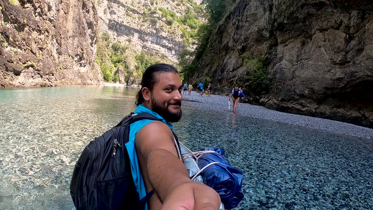Young man with travel backpack filming himself walking on shallow cold water of mountain river