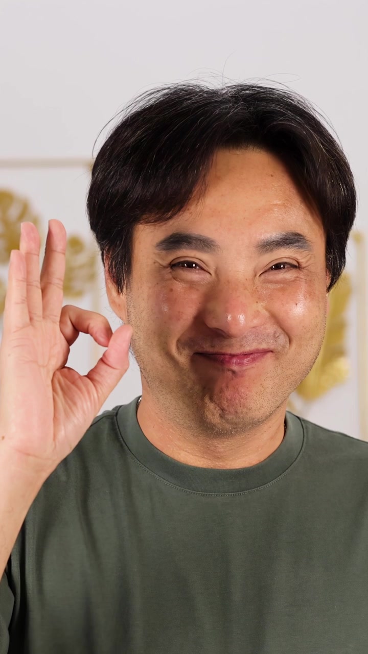 Uomo asiatico sorridente in camicia verde che fa il gesto dell'OK in interni con uno sfondo morbido e neutro