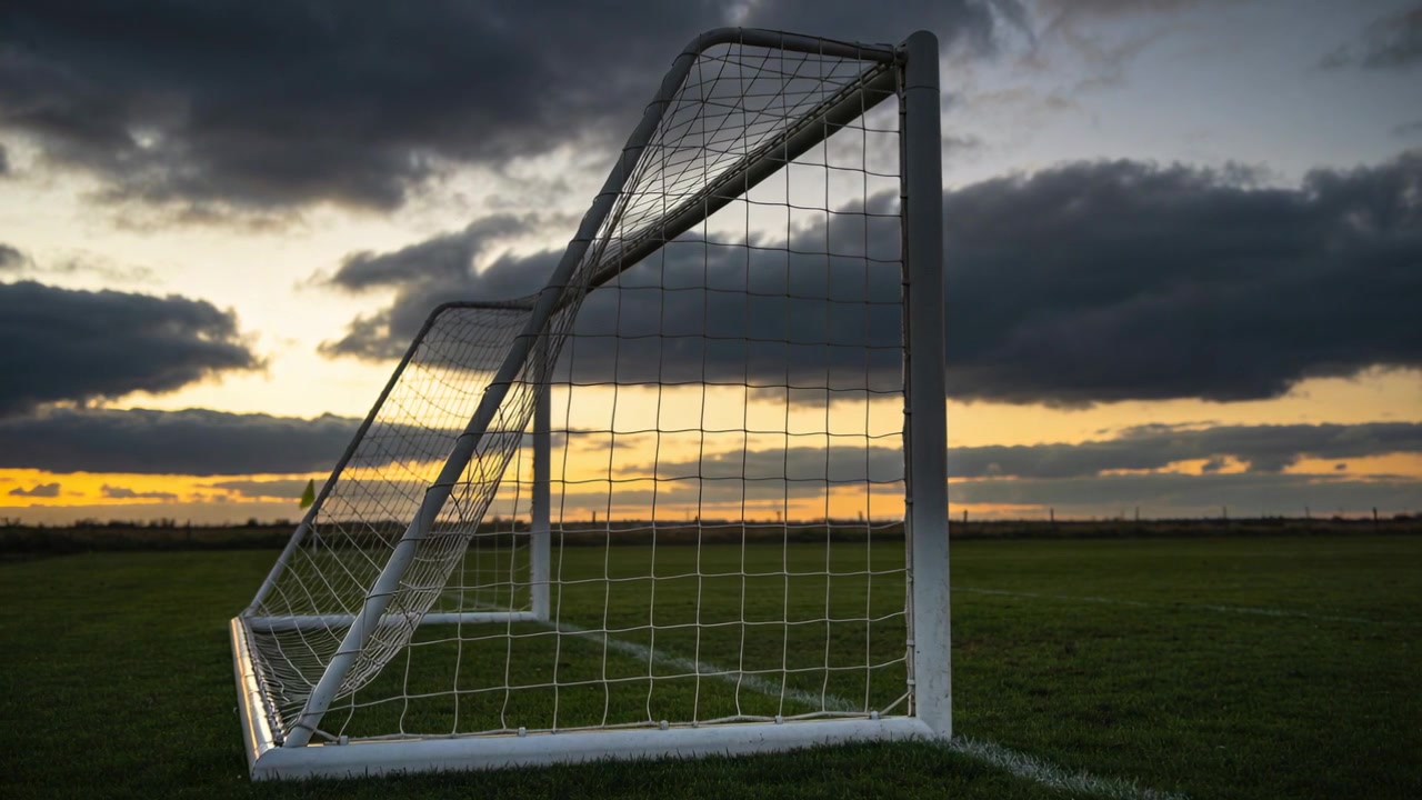 Showing white soccer goal net shifting as breeze blowing and sun sinking on grassy pitch, clouds