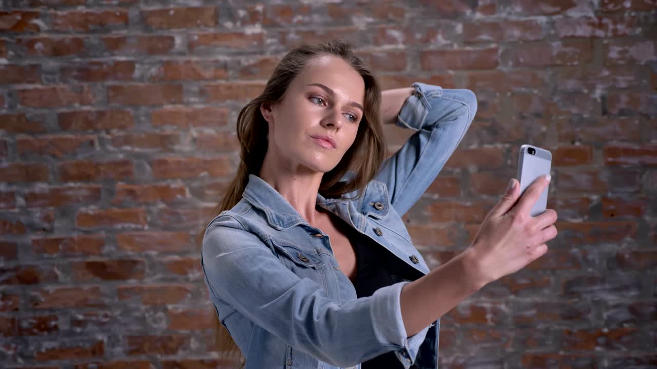 una joven linda está haciendo selfie en su teléfono inteligente, telón de fondo de ladrillo, concepto de comunicación