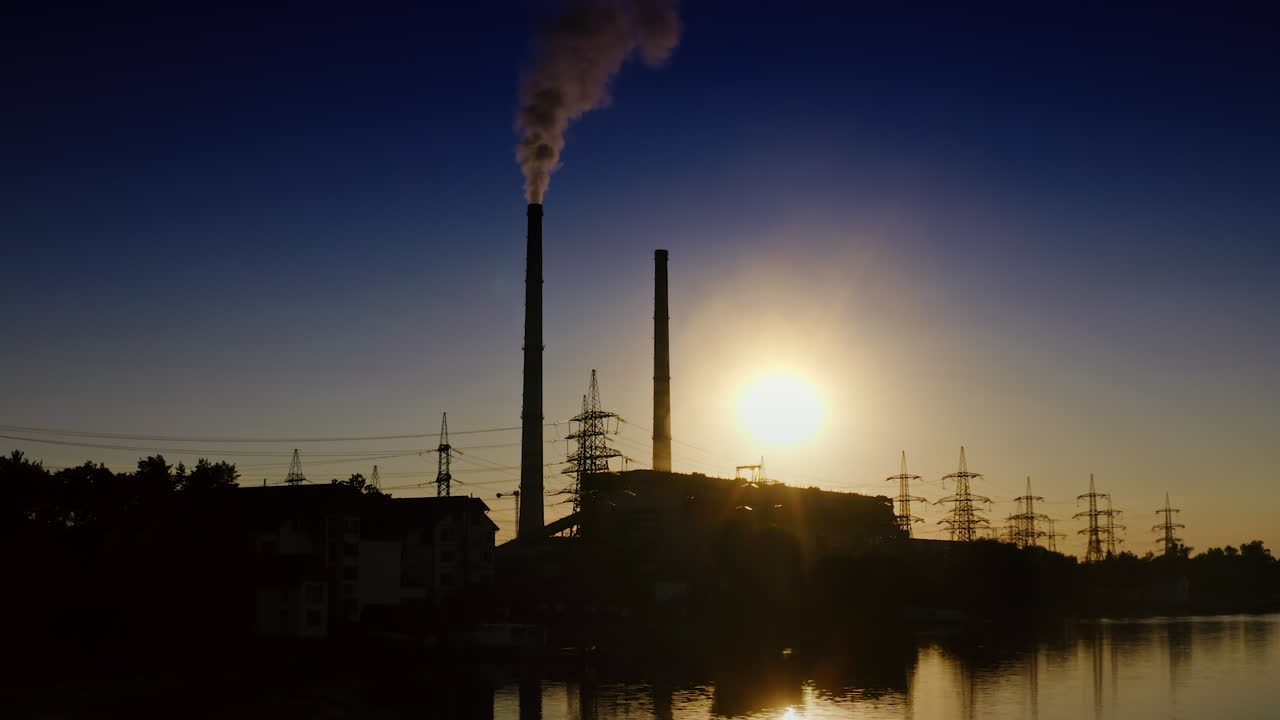 Silhouette of factory at sunset. Dark smoke from industrial pipe filling the air. Harmful manufacturing near the river in the evening. Ecological disaster.