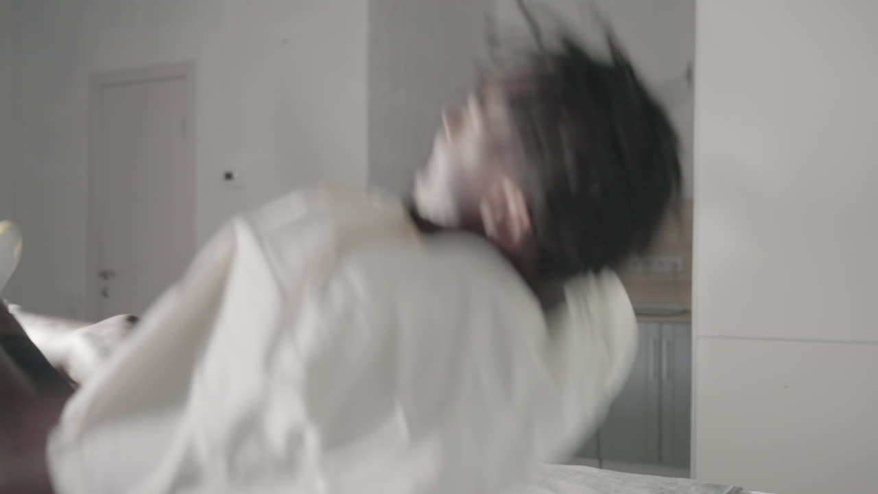Man in white t-shirt bending down to pick up backpack in minimalist bedroom, with green headboard, bed with neatly folded towels, and clean modern decor in the background