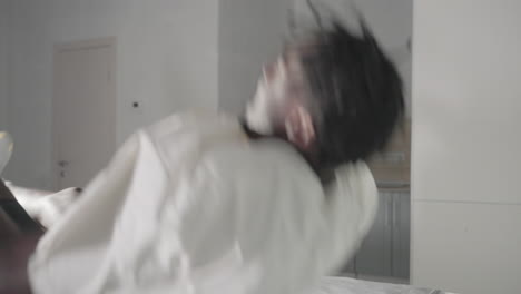 man in white t-shirt bending down to pick up backpack in minimalist bedroom, with green headboard, bed with neatly folded towels, and clean modern decor in the background
