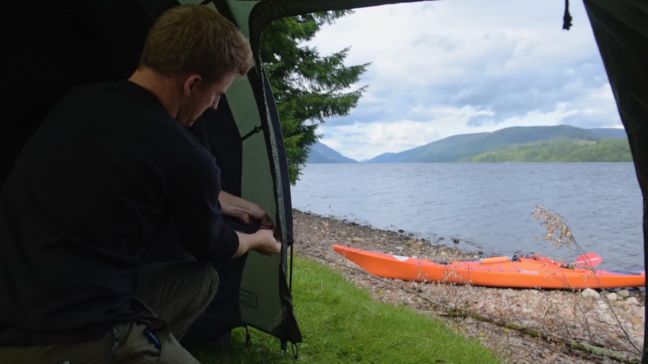 Man Setting Up The Interior Of A Tent Near Caledonian Canal In Scotland With Orange Kayak On The Shore - Medium Shot