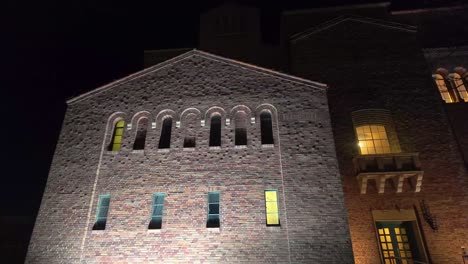 footage looking up at illuminated vintage brick building with rows of arched windows glowing yellow from within at night