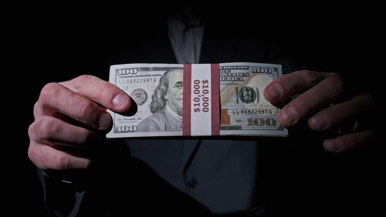 un hombre de negocios en traje muestra una pila de 10.000 dólares estadounidenses en fondo negro.