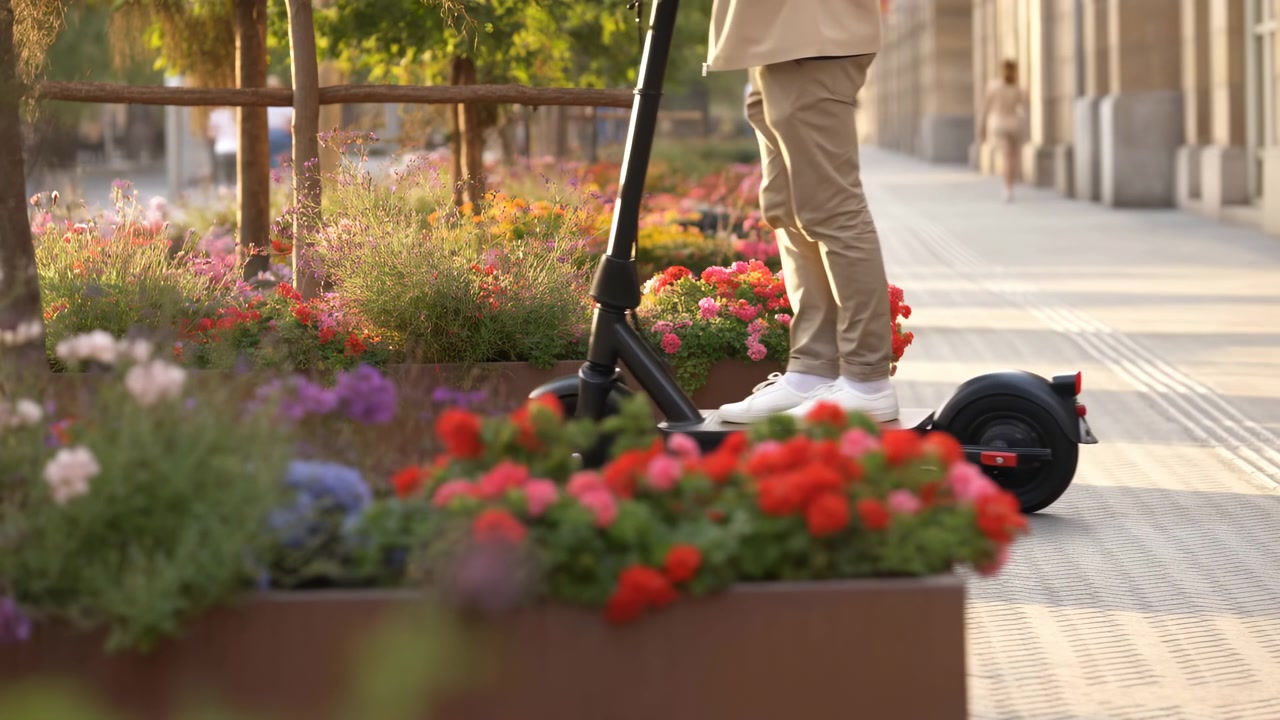 Parte inferiore del corpo di una persona ferma su uno scooter elettrico accanto a una fila diagonale di fioriere rialzate color ruggine, straripanti di fiori rossi, rosa, arancioni, viola e bianchi, vegetazione e luce solare calda