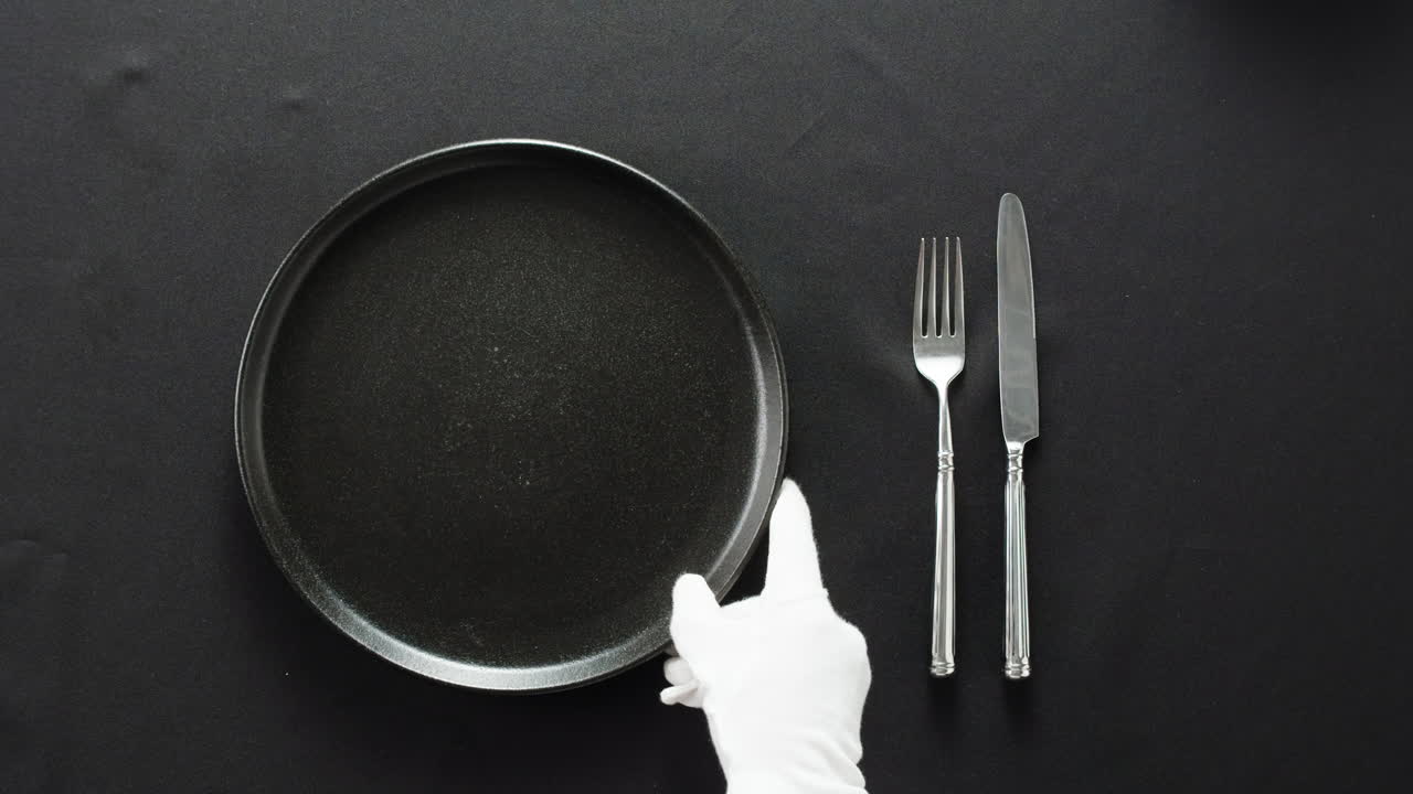 Minimalist table setting with white plate, fork, and knife arranged neatly, and a gloved hand adjusting cutlery.