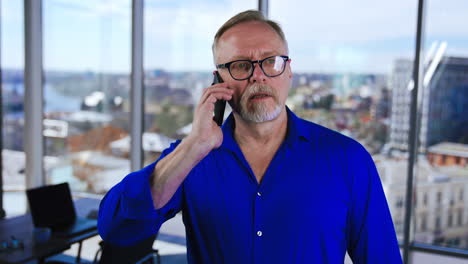 serious grey-haired man wearing glasses speaks on the phone in the office. business talk on the smartphone at work.