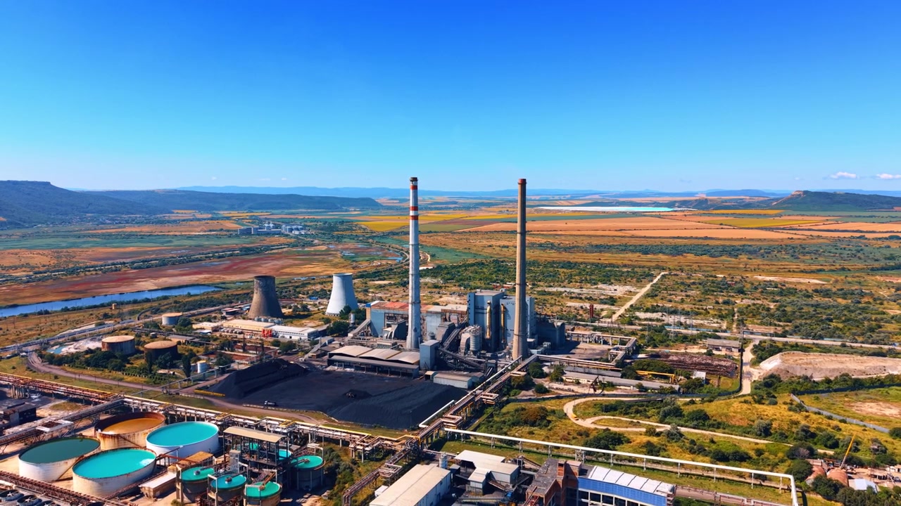 Ampla vista aérea de uma usina de energia com chaminés altas. Duas chaminés altas e torres de resfriamento ficam perto de bacias de água em uma paisagem aberta sob céu azul.