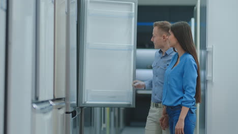 a young married couple open the door of the new refrigerator inspect the design and quality before buying in a consumer electronics store. read the information on the price tag and characteristics