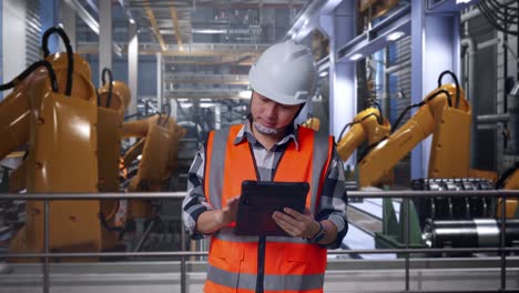 asian male engineer with safety helmet working on a tablet while standing with industrial robotic arms working