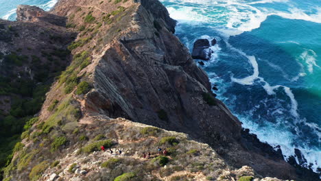Aerial-top-down-of-hiker-group-walking-downhill-the-steep-cliff-mountain-during-agitated-ocean-water-in-background
