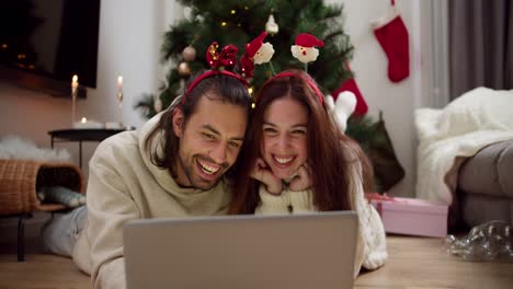 Happy-couple-guy-and-girl-in-New-Year's-clothes-lie-on-the-floor-in-front-of-a-gray-laptop-watching-a-Christmas-movie-near-the-Christmas-tree-in-a-cozy-decorated-room