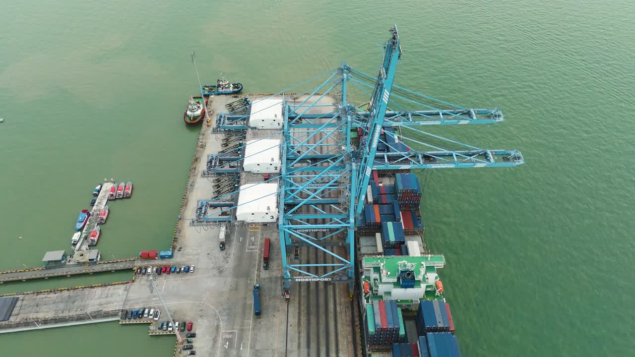 Top-down aerial shot of massive cargo cranes at Northport in Klang, Malaysia, loading containers onto ships amid busy port activity and green coastal waters.