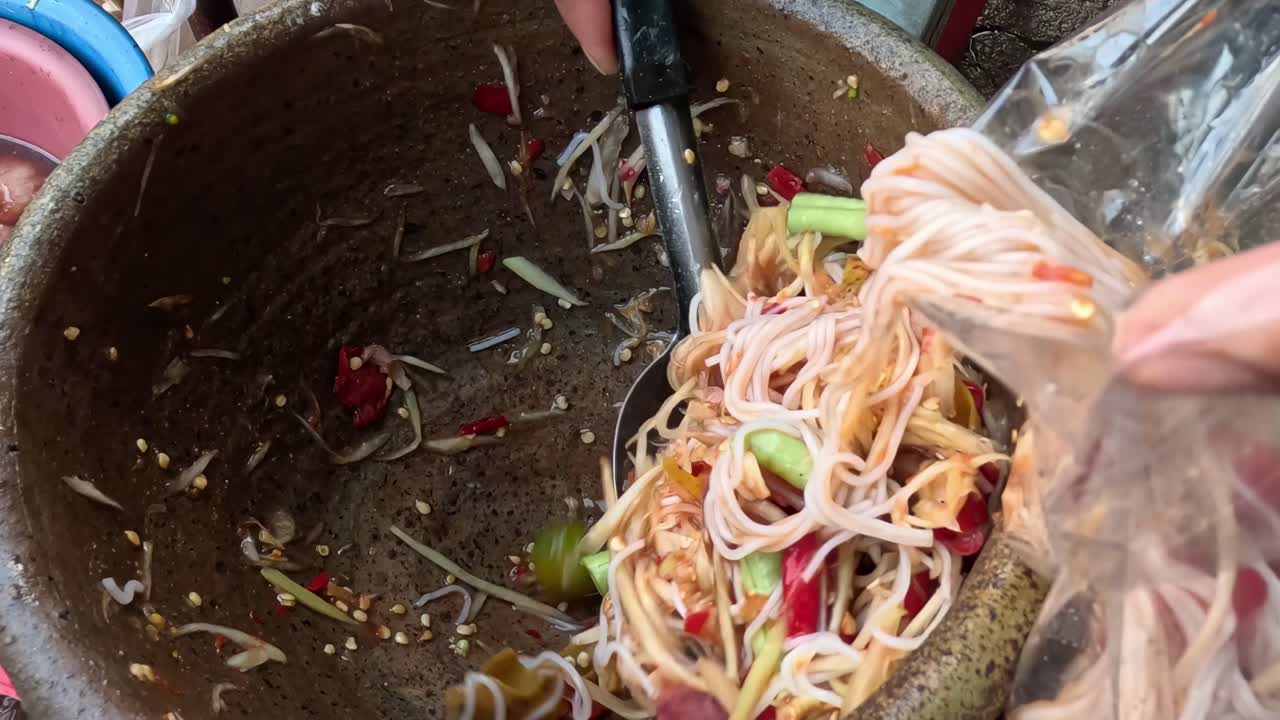 A close-up view of noodles and vegetables being scooped into a plastic bag from a mortar.