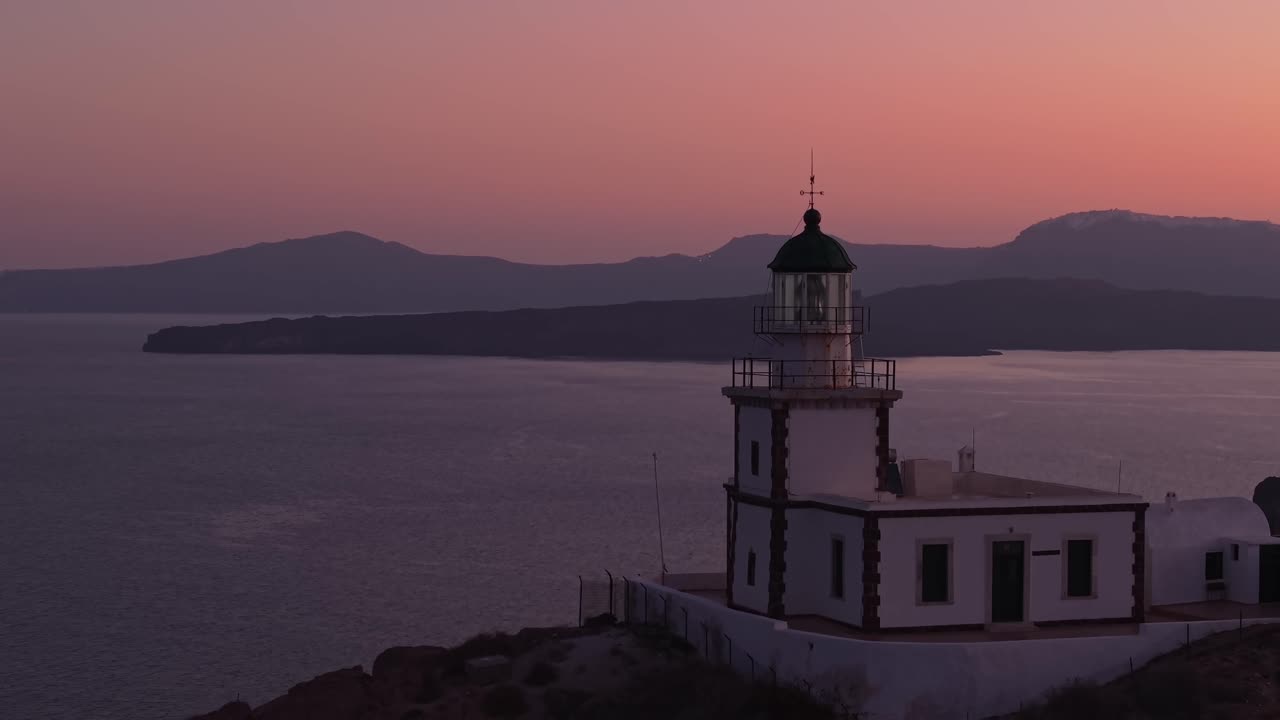 Aerial drone footage revealing the Akrotiri Lighthouse in Santorini at sunrise, with a soft pink sky and the volcano visible in the background — a peaceful Mediterranean scene