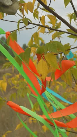 parts of animal skull hang on tree with green and yellow leaves. multicolored ribbons flutter in wind tied to cow bone horn and branches on autumn lawn under blue sky