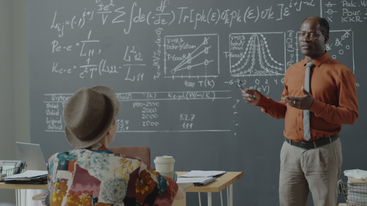 A professor lectures students in a classroom with a blackboard full of complex equations and graphs