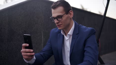 Hombre-De-Negocios-Emocionado-Y-Sonriente-Sentado-En-Las-Escaleras-Y-Hace-Video-Cahtting-En-El-Teléfono-Inteligente-Afuera