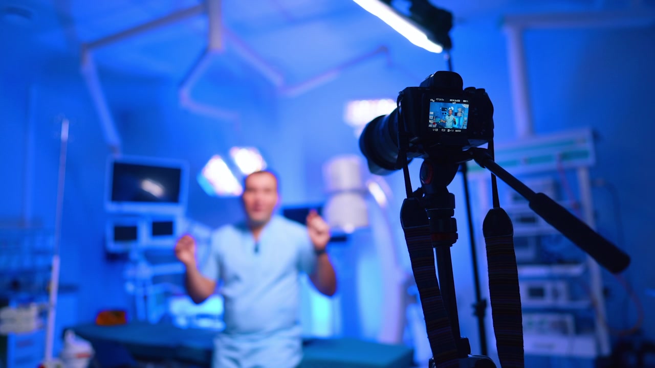 Telecamera che riprende un medico in piedi in una sala operatoria chirurgica blu. L'attrezzatura su treppiede con anteprima posteriore mostra un presentatore sfocato in camice chirurgico tra lampade chirurgiche e monitor.