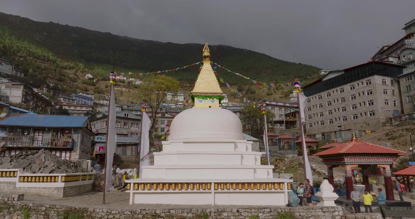 Namche Bazaar, Nepal, luftfoto med drone som zoomer inn og nærmer seg en hvit buddhistisk chorten stupa med tradisjonelle hus og frodige grønne åser i bakgrunnen.