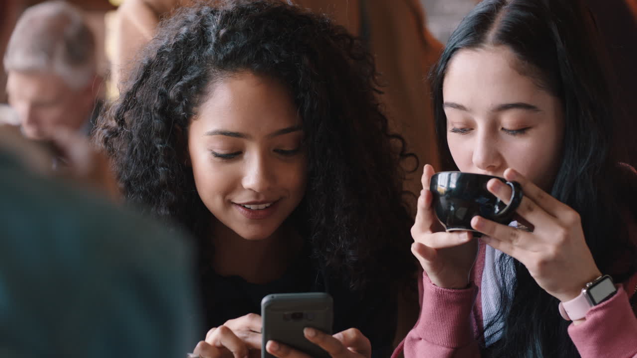 happy teenage girl friends using smartphones drinking coffee in cafe texting hanging out sharing gossip enjoying chatting together