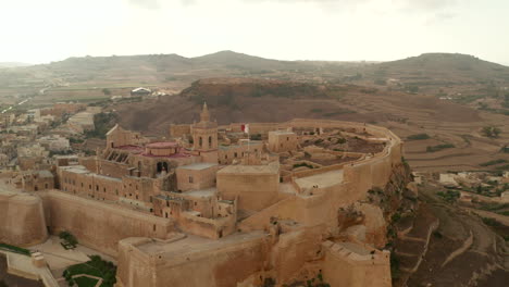 Establecimiento-De-Una-Toma-Del-Fuerte-Del-Castillo-De-Gozo-Con-Un-Castillo-Ondeando-La-Bandera-De-Malta-En-Un-Hermoso-Color-Marrón-Arena,-Tobogán-Aéreo-A-La-Derecha