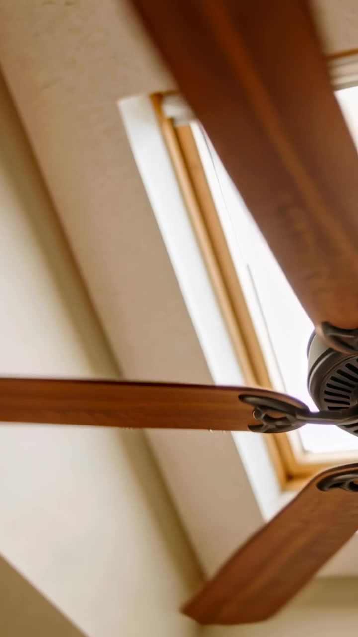 Video verticale: Motore che alimenta un ventilatore in legno a tre pale che gira sotto un lucernario in una stanza con una tenda.