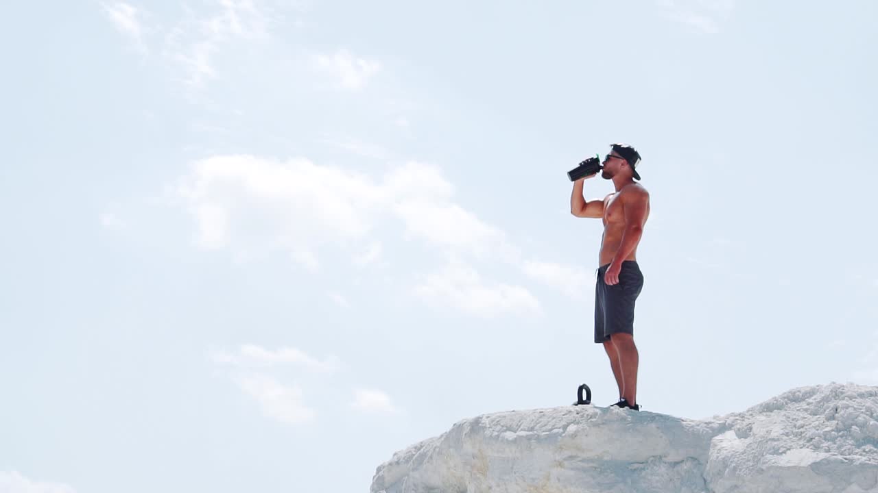 An athletic man drinks an energy drink from a bottle after training outdoors. Slow motion