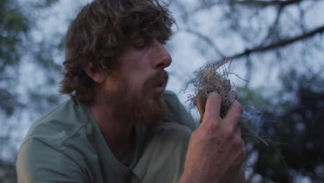 bearded caucasian male survivalist blowing on smoking tinder to start fire at camp in wilderness