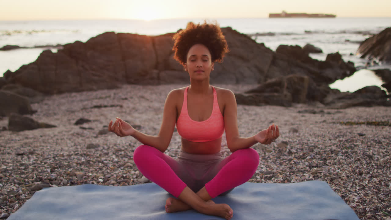 Gün batımında denizin yakınındaki kayaların üzerinde yoga ve meditasyon yapan Afrikalı Amerikalı bir kadın.