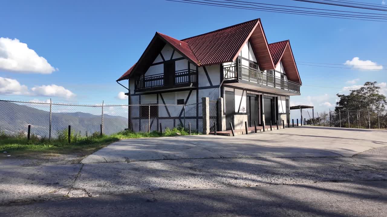 Rumah gaya Alpen dengan atap merah yang terletak di titik pandang gunung. Pemandangan indah dengan langit biru, pegunungan jauh, dan latar depan pinggir jalan di lingkungan dataran tinggi pedesaan.