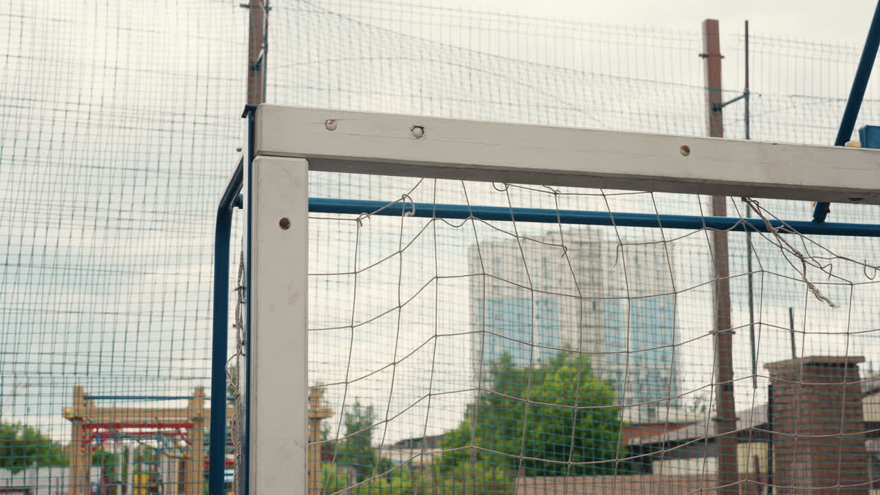 urban football goalpost under rainy sky, wet net glistening with droplets, industrial skyline blurred in background, empty pitch framed by metal posts and mesh fence, overcast light creating cinematic