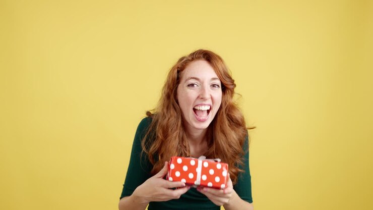 Happy Redhead Woman Showing Excitement and Holding a Gift Box on Yellow Background