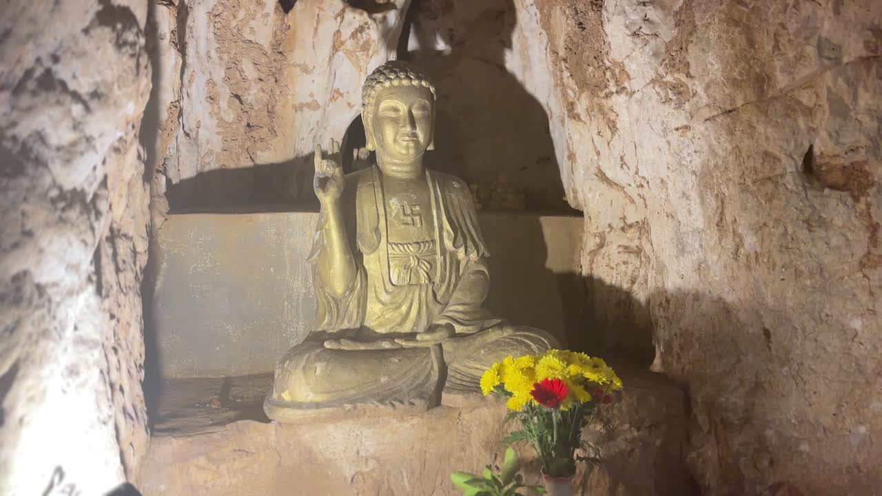 Statua di Buddha seduta in un santuario rupestre all'interno delle Montagne di Marmo Ngu Hanh Son a Da Nang con luce calda, pareti di pietra grezza, piccolo altare con fiori e un'atmosfera spirituale pacifica