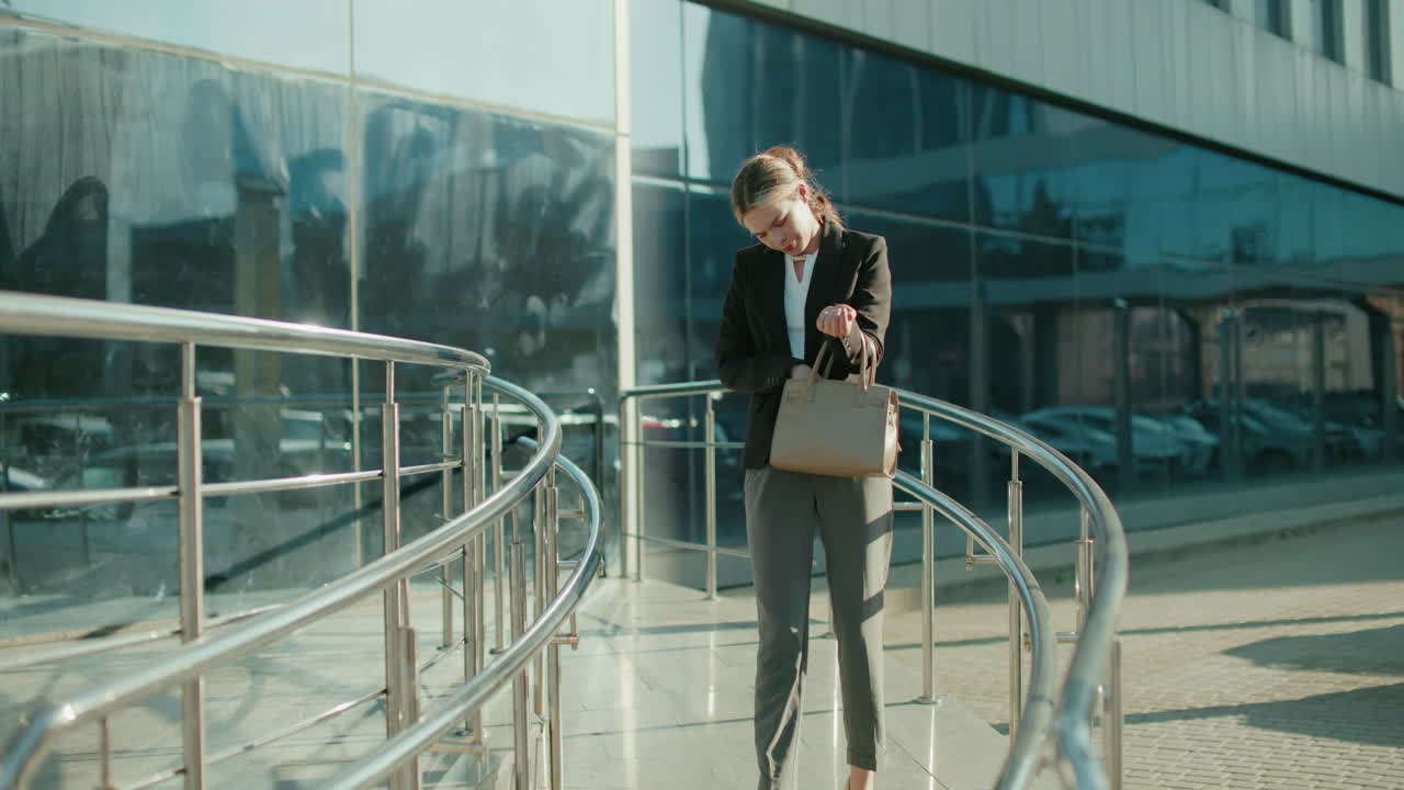 Professional woman walking down office ramp, contemplatively checking handbag to retrieve car key, surrounded by sleek metal railings, glass building reflecting urban setting