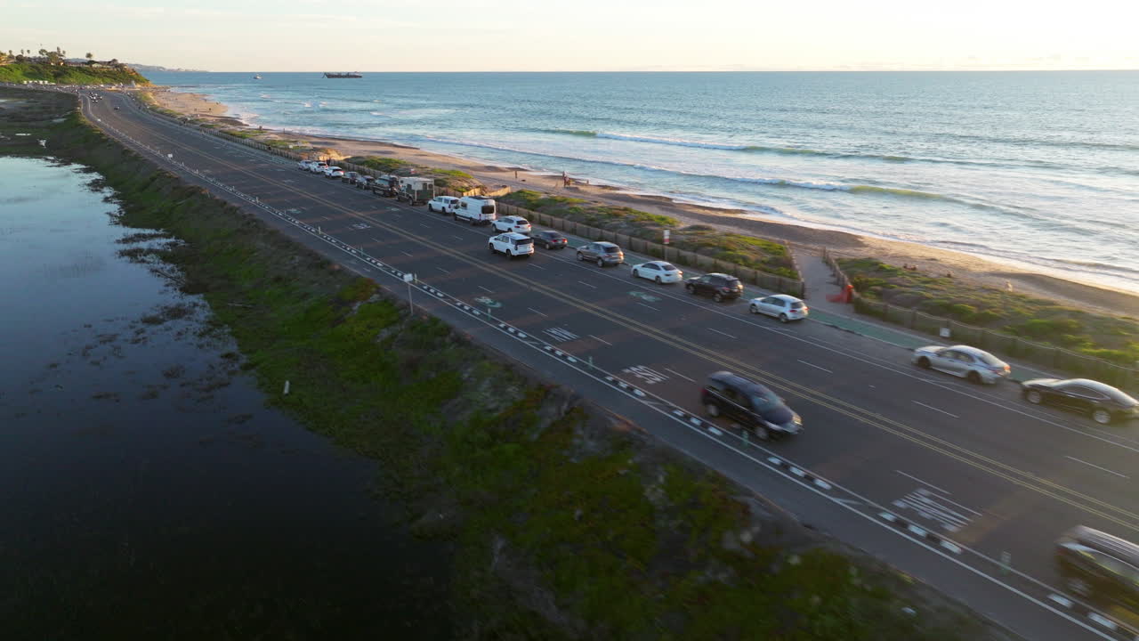 White SUV Driving Along The Coastal Highway Lined With Cars Beside The Ocean At Sunset In Del Mar, California. - aerial cinematic shot